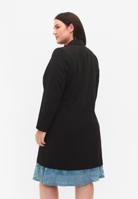 Zwarte op maat gemaakte jas met lange mouwen, met een gladde textuur en gestructureerd ontwerp. Gelaagd over een lichtblauwe kanten jurk.