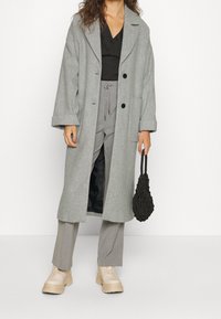 Vrouw in een lange grijze jas over een grijze broek en een zwart top, met een zwart netten handtas en beige schoenen met dikke zolen.