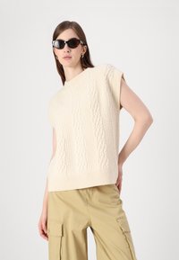 Pleteni pulover u krem boji s pletenim uzorkom, kratkim rukavima i rebrastim rubom, u kombinaciji s khaki cargo hlačama. Model nosi crne sunčane naočale u obliku mačjih očiju.