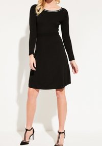 Robe noire à manches longues, corsage ajusté et jupe évasée. Présente une accentuation blanche le long de l'encolure. Portée avec des talons noirs.