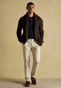 Brūns adīts šalle ar apkakli virs jūras zila un bordo svītrainā polo krekla. Krēmkrāsas bikses un brūnas mokasīnu kurpes noslēdz apģērbu.