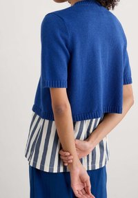 Blue cropped knit cardigan with short sleeves, ribbed edges, worn over a striped blouse in white and navy, paired with blue trousers.