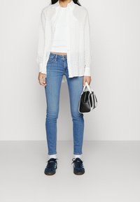 Bílá košile na knoflíky, fitted bílý top bez rukávů, modré úzké džíny, námořnické semišové tenisky, nese malou černobílou kabelku.