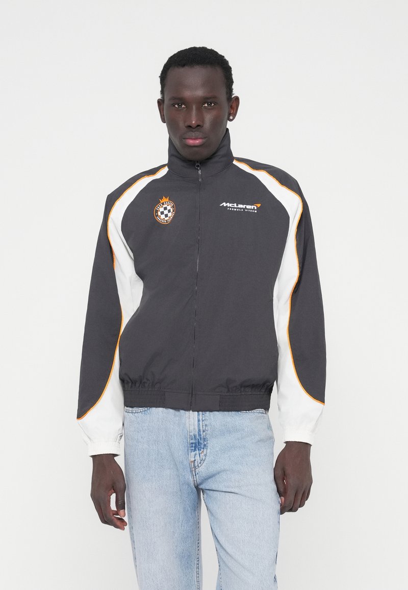 Μπουφάν McLaren σε μαύρο χρώμα με λευκές και πορτοκαλί λεπτομέρειες, διαθέτει ψηλό γιακά, φερμουάρ μπροστά και ελαστική μέση. Λεπτομέρεια με κεντημένο λογότυπο.