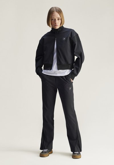 Schwarze cropped Zip-Jacke mit hohem Kragen, kombiniert mit schwarzen Hosen mit seitlichen Schlitzen und Logo-Details. Getragen über einem weißen Shirt.
