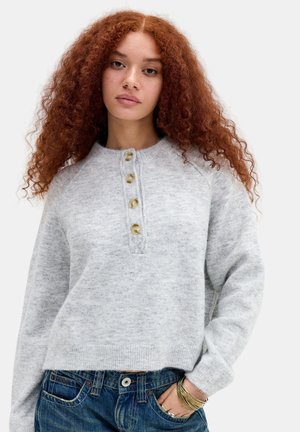 Ung kvinde med langt krøllet rødt hår iført en lysegrå cardigan med knapper og blå jeans med armbånd på håndleddet.