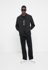 Sort button-up jakke med struktureret overflade, parret med en sort grafisk t-shirt, sorte jeans og hvide sneakers. Model bærer sorte solbriller.