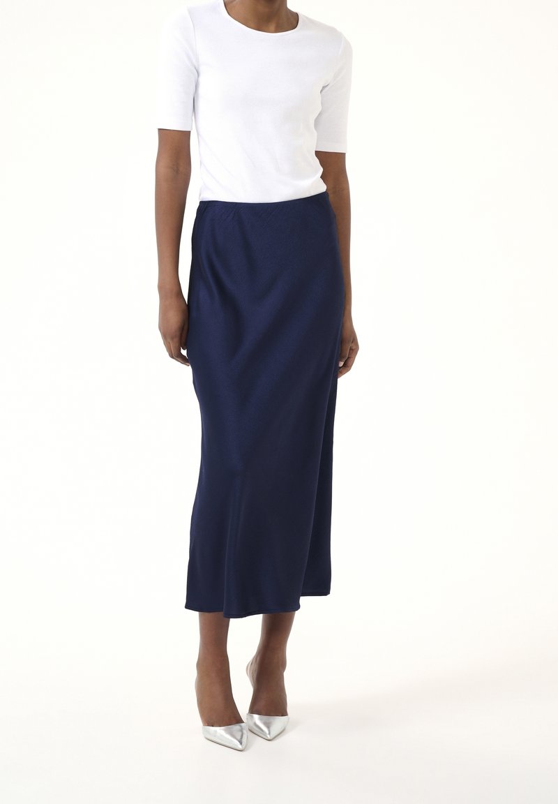 Jupe midi bleu marine en tissu texturé, associée à un haut blanc à manches courtes et des escarpins argentés à bout pointu. Silhouette lisse.