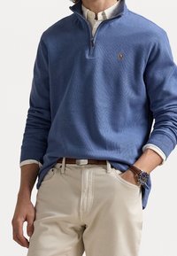 Suéter azul de manga larga con cuello de cremallera de un cuarto, hecho de una tela texturizada; presenta un pequeño logo y se combina con pantalones claros.