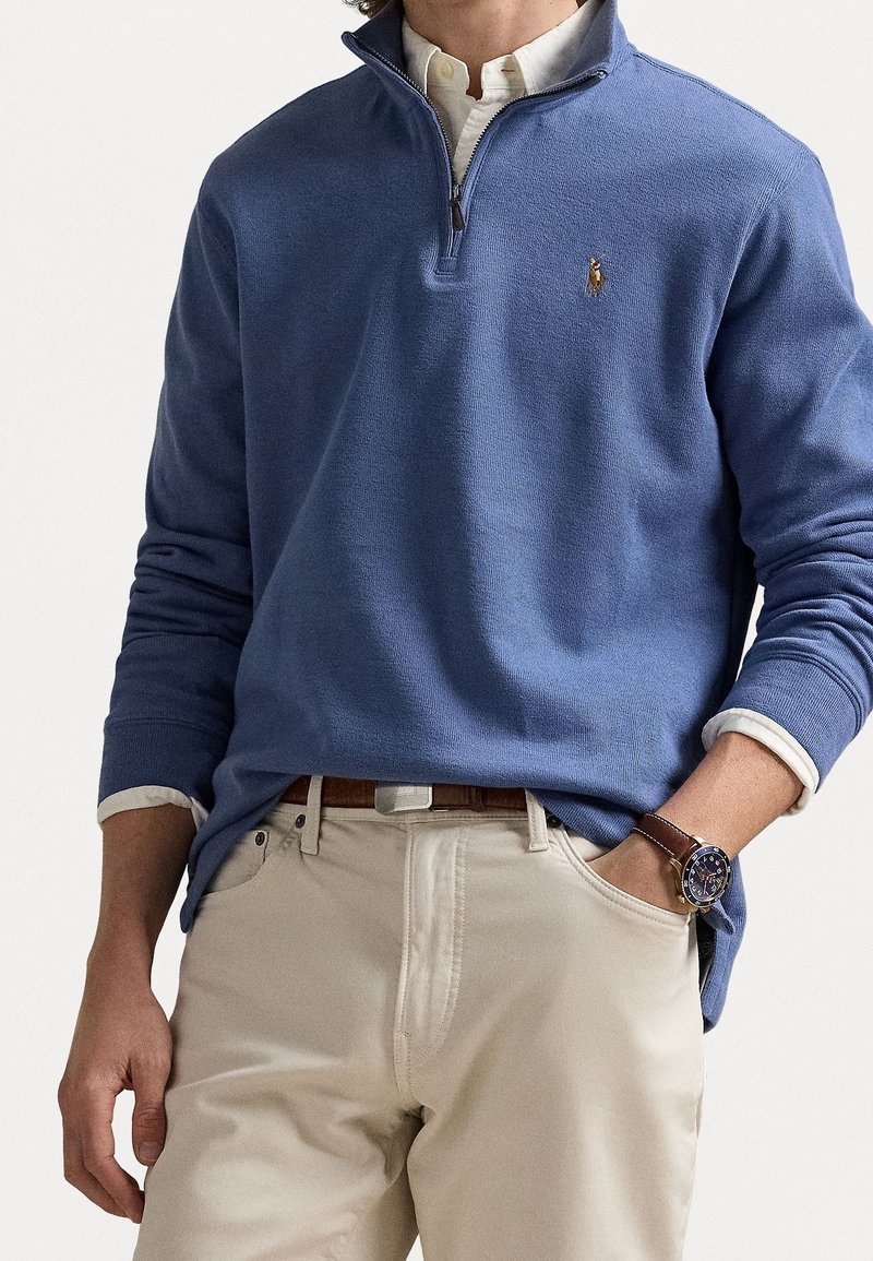 Suéter azul de manga larga con cuello de cremallera de un cuarto, hecho de una tela texturizada; presenta un pequeño logo y se combina con pantalones claros.