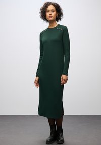 Abito lungo a maniche lunghe di colore verde scuro con texture a costine, spacchi laterali e bottoni decorativi sugli spallacci. Indossato con stivaletti neri sopra la caviglia.