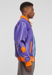 Kožená bomber bunda v purpurové barvě s oranžovými akcenty, se hvězdicovými nášivkami a žebrovaným pasem. Zapínání na zip.