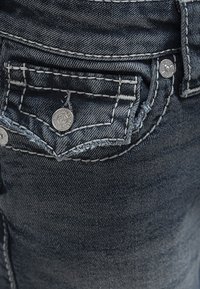 Gros plan sur la poche d'un jean en denim bleu foncé avec des coutures blanches détaillées et des boutons-pression en métal.