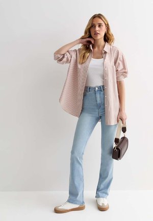 Camisa de botones a rayas en rosa claro y blanco con mangas remangadas, combinada con jeans de talle alto en azul claro y zapatillas blancas. Se sostiene un bolso marrón.