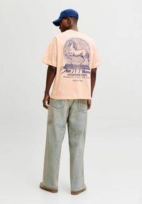 Persoană purtând un tricou piersică cu un grafic de cal și text, blugi decolorați deschiși, pantofi de culoare cărămizie și o șapcă de baseball albastră, stând cu fața spre spate.