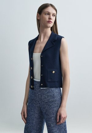 Kobieta ubrana w granatową bezrękawnikową kamizelkę na guziki, biały top na ramiączkach oraz niebieskie teksturowane spodnie o szerokich nogawkach, stojąca na jednolitym tle.