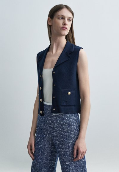 Femme portant un gilet sans manches bleu marine boutonné, un débardeur blanc et un pantalon large bleu texturé, debout devant un fond uni.