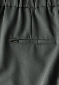 Tissu gris foncé avec une texture lisse, comportant une poche passepoilée horizontale et une petite étiquette en métal. Détail de ceinture froncée visible.