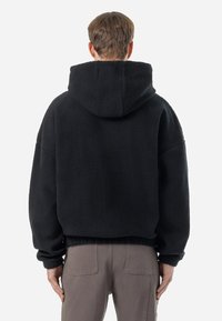 Haut de survêtement en polaire noire avec un col haut, coupe oversize et ourlet côtelé. Présente une surface texturée et une capuche réglable.