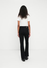 Kvinde med krøllet hår iført en hvid kortærmet top, sorte bootcut bukser og sorte platfodede sko, som står vendt mod en hvid væg.
