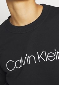 Felpa nera con colletto a girocollo, polsini a coste e un grande logo bianco "Calvin Klein" sul petto. Texture liscia.