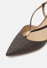 Chaussure plate marron à bout pointu avec motif, bride en T et détail de boucle circulaire dorée sur semelle intérieure beige sur fond blanc.