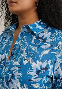 Blå blommig skjorta med stora vita och marinblå blommor, knäppt look, och krage; tillverkad av lätt material med en mjuk textur.