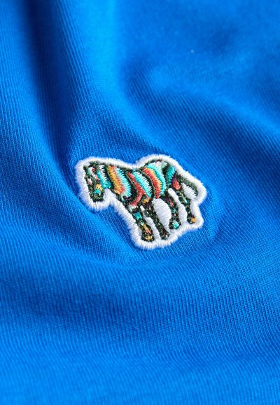 Blå stof med en broderet, stiliseret zebra-design med multikoloriske striber, omridset i hvidt, på en struktureret overflade.