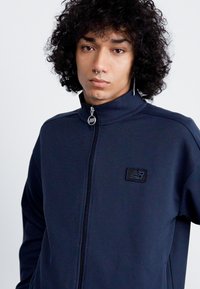 Navy zip-up jacket with high collar, textured fabric, and a black New Balance logo on the left chest. Features a silver zipper.