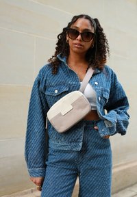 Giubbotto di denim e jeans abbinati con una texture a motivi; il modello tiene una borsa a tracolla beige e crema con zip e tracolla.