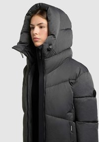 Giacca puffer imbottita di colore grigio scuro, con colletto alto, grande cappuccio, chiusura con zip e dettagli di cucitura evidenti.