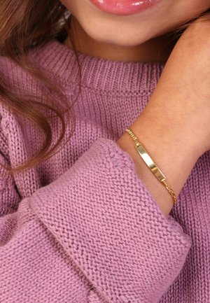 Gouden armband met een glad, plat plaatje, schakelketting ontwerp, gedragen aan een hand die rust tegen een lichtpaarse gebreide trui.