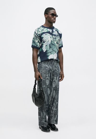 Cămașă tricotată albastru marin cu model floral verde, asortată cu pantaloni negri largi cu model. Accesoriile includ o geantă neagră și ochelari de soare.