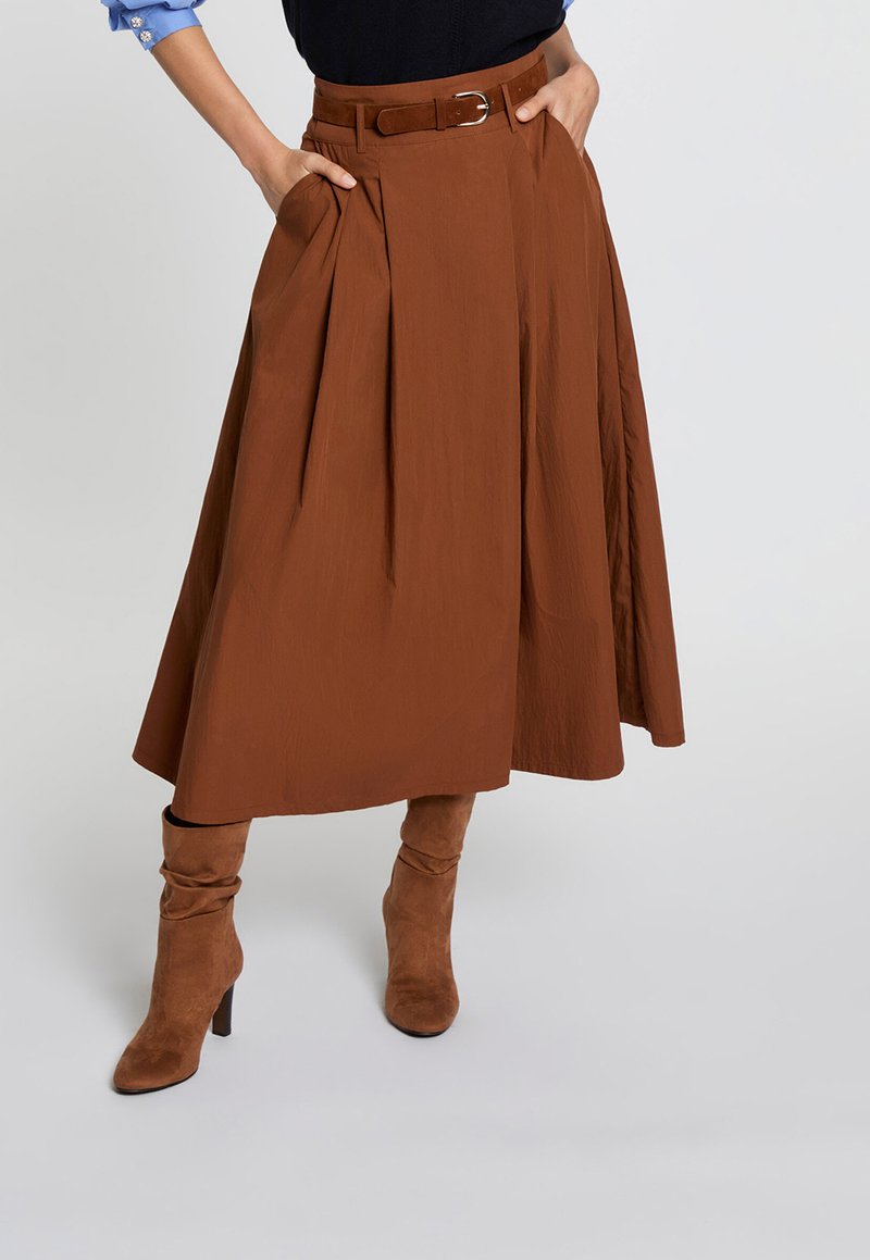 Woman wearing a brown high-waisted midi skirt with belt and matching suede heeled boots, hands in skirt pockets against a light background.