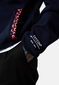 Manche de sweat-shirt bleu foncé avec le texte blanc "ESTEEM" et une bordure en imprimé léopard rouge sur la poche, main partiellement dans la poche d'un pantalon noir montrant un poignet tatoué.