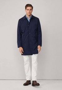 Capa de chuva azul-marinho com colarinho, fecho de botão único e bolsos laterais, usada sobre uma camisa azul-clara e calças brancas, combinando com sapatos castanhos.