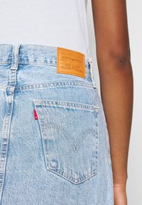 Poche de jean denim bleu clair avec une étiquette de marque en cuir Levi's et une étiquette en tissu rouge, portée avec une chemise blanche sur un bras à la peau foncée.