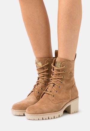 Béžové semíčkové kotníkové boty na šněrování s blokovým podpatkem, vybavené kovovými očky, embosovaným logem a texturovanou gumovou podrážkou.