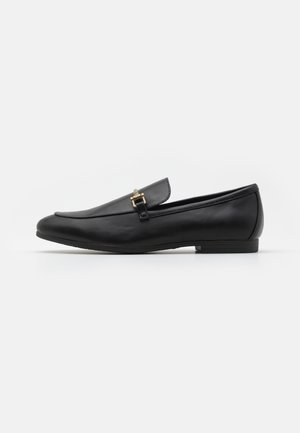 Mocassin en cuir noir sans lacets avec un petit talon et une petite boucle en métal doré sur le dessus.
