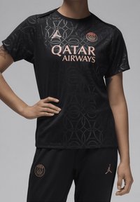 Camiseta deportiva negra con un patrón circular texturizado, que presenta un logo rosa de Qatar Airways y PSG, complementada por pantalones negros con detalles en dorado.