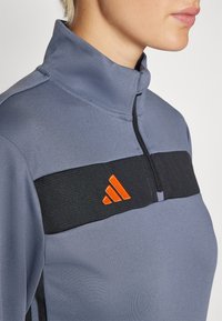 Sivi pulover s visokim ovratnikom i kontrastnim crnim pojasom. Ima narančasti logo i zatvaranje na patentni zatvarač, napravljen od teksturiranog materijala.