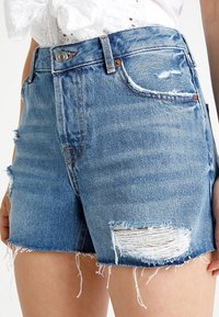 Džíny šortky v světle modré barvě s rozpáraným lemem, vysokým pasem a opotřebovanými detaily, včetně roztrhaných záplat a vybledlých míst.
