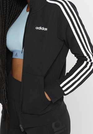 Kobieta ubrana w czarną kurtkę Adidas zapinaną na zamek z białymi paskami na rękawach, jasnoniebieski sportowy biustonosz i czarne legginsy, ręka w kieszeni kurtki.