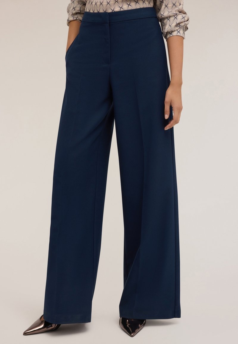 Femme portant un pantalon bleu marine taille haute à jambes larges avec poches latérales et chaussures pointues métalliques, debout devant un fond uni.
