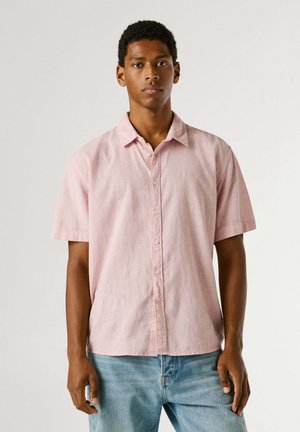 Jonge man die een lichtroze kortärmlig overhemd en een blauwe spijkerbroek draagt, staand tegen een effen lichte achtergrond.