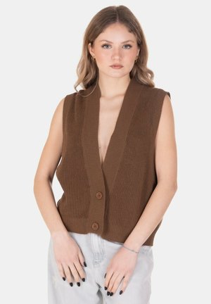 Donna con cardigan marrone lavorato a maglia senza maniche con due bottoni sopra jeans chiari in denim, in piedi contro sfondo bianco.