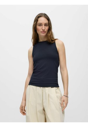 Femme portant un haut noir sans manches côtelé et un pantalon ample beige, debout devant un fond gris clair uni.