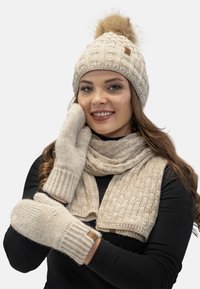Beige pletená čepice s bambulí, odpovídající šála a rukavice. Texturovaný vzor a vroubkované manžety, vše vyrobeno z měkkého, teplého materiálu.