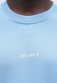 Közeli kép egy személyről, aki világoskék kerek nyakú pulóvert visel, amelyen fehérrel van ráírva a mellkason, hogy "LES DEUX".