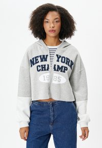 Gekürzter grauer Kapuzenpullover mit dunkelblauem Text "NEW YORK CHAMP" und Jahr "1985." Weiße Ärmel, Frontreißverschluss, lässige Passform, weicher Stoff.